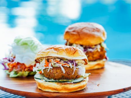 The image shows two burgers topped with coleslaw and pickles on a wooden serving board, with a pool in the background.