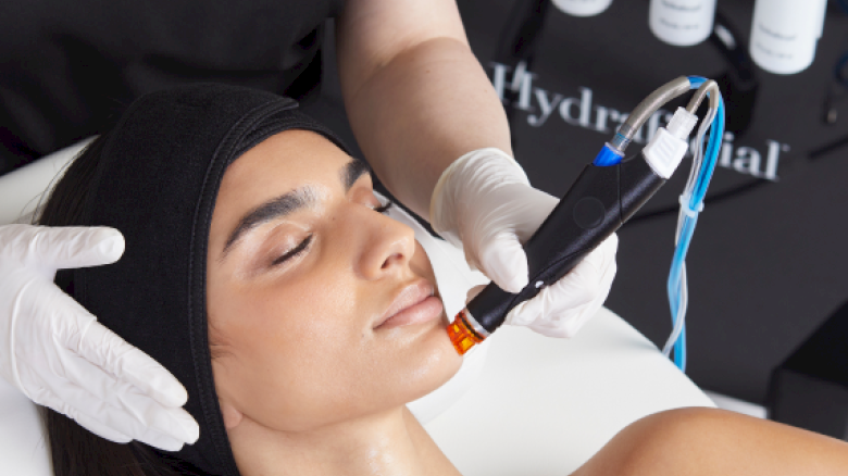 A woman is receiving a HydraFacial treatment at a spa. An aesthetician is using a device on her face while she relaxes with eyes closed.