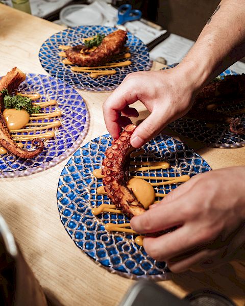 A person is arranging food on a blue patterned plate, garnished with sauces and herbs. Similar plates with food are visible in the background.