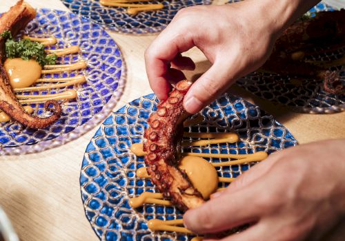 A person is arranging food on a blue patterned plate, garnished with sauces and herbs. Similar plates with food are visible in the background.