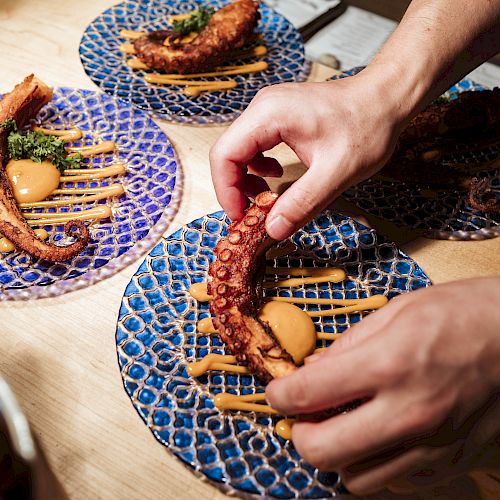 A person is arranging food on a blue patterned plate, garnished with sauces and herbs. Similar plates with food are visible in the background.