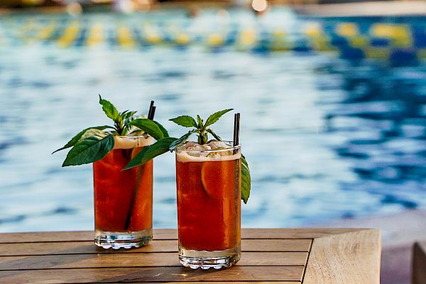 Two refreshing drinks with garnishes on a wooden table, situated poolside with lounge chairs and greenery in the background.