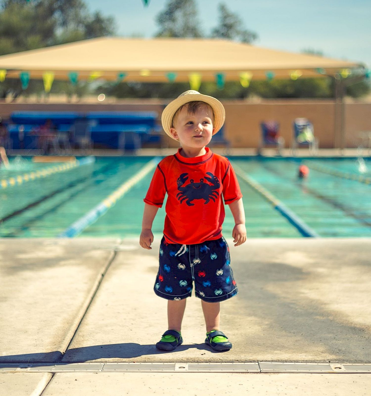 A young child in a red shirt and hat stands confidently by a swimming pool, with people and greenery in the background.