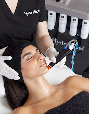 A person is receiving a HydraFacial treatment from a professional wearing gloves. The person lies down with closed eyes during the procedure.