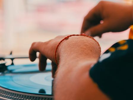 A person is operating a turntable, likely DJing, with a blurred background. They wear a bracelet on their left wrist, while adjusting the record.