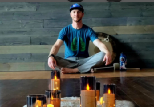 A person sits cross-legged on a wooden floor with a fuzzy rug and multiple lit candles in glass holders, creating a calming ambiance.