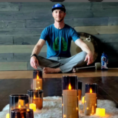 A person sits cross-legged on a wooden floor with a fuzzy rug and multiple lit candles in glass holders, creating a calming ambiance.