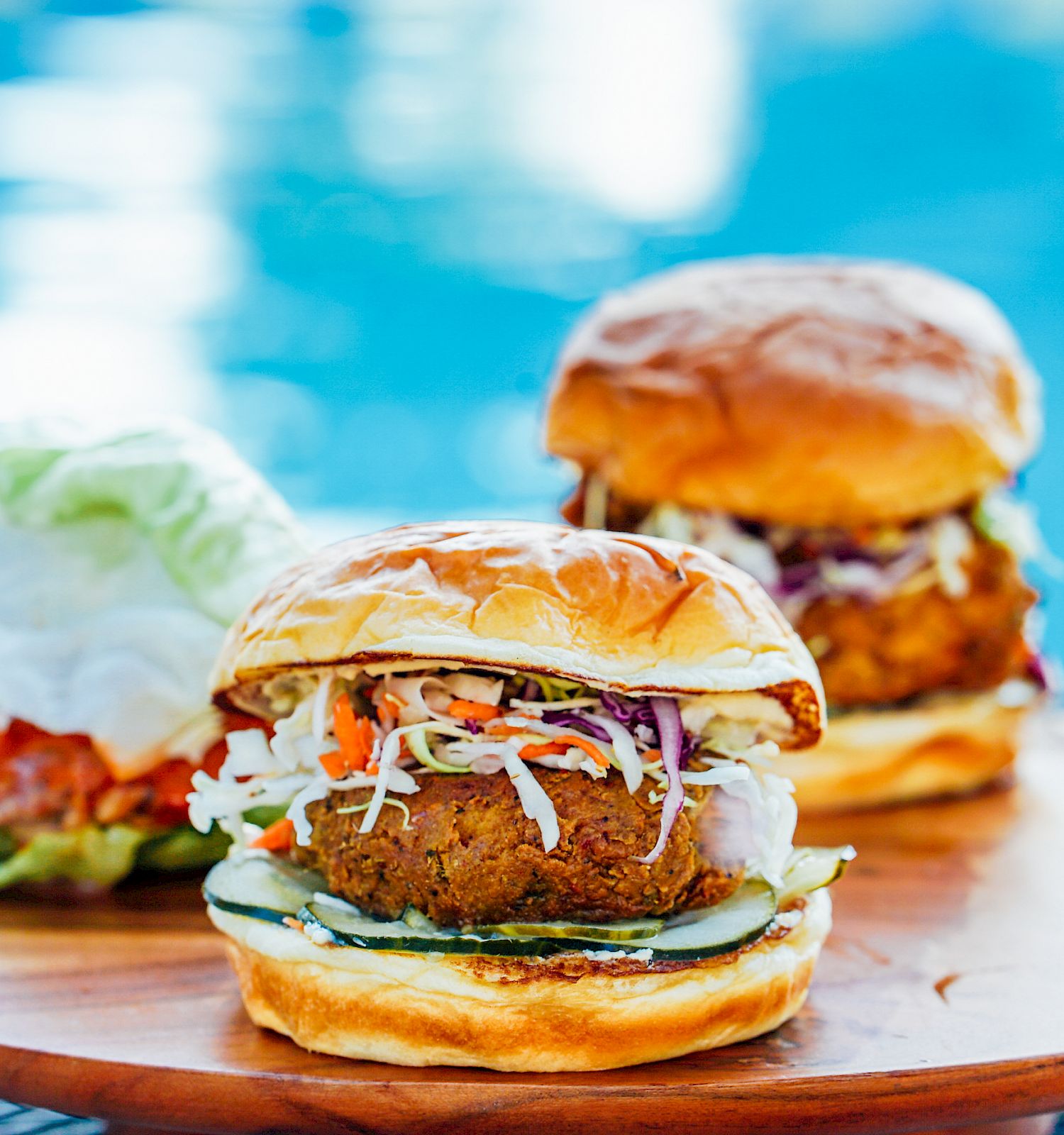 Two fried chicken sandwiches with coleslaw and cucumber on a wooden board, near a pool in the background, with a side salad.