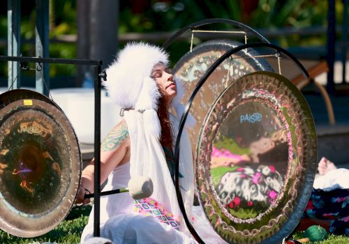 A person wearing a white headdress and white outfit plays large gongs outdoors, surrounded by greenery and sunlight.