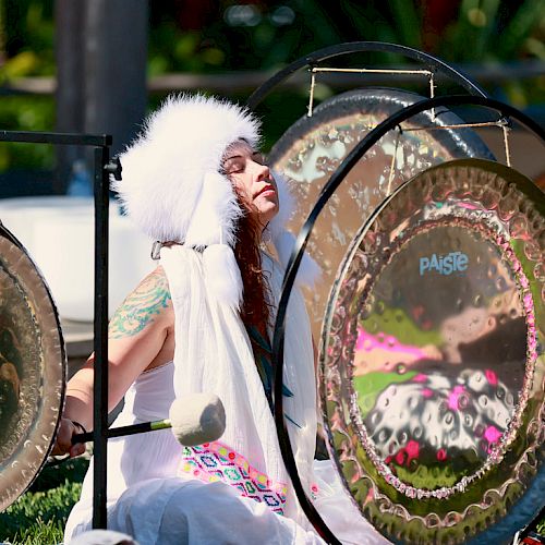 A person wearing a white headdress and white outfit plays large gongs outdoors, surrounded by greenery and sunlight.