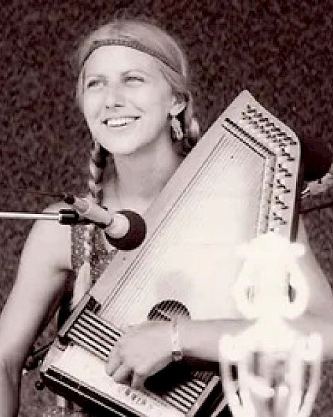 A person is playing an autoharp and smiling while standing in front of a microphone, with a blurred background in the image.