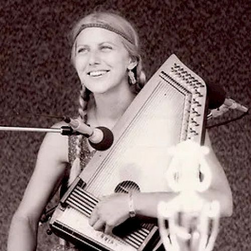 A person is playing an autoharp and smiling while standing in front of a microphone, with a blurred background in the image.