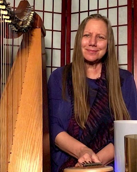 A person with long hair sits among musical instruments, including a harp and several other items, in front of a folding screen.