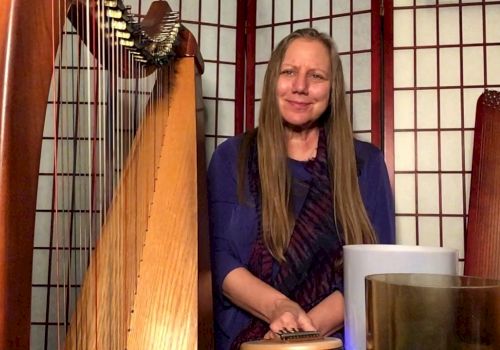 A person with long hair sits among musical instruments, including a harp and several other items, in front of a folding screen.