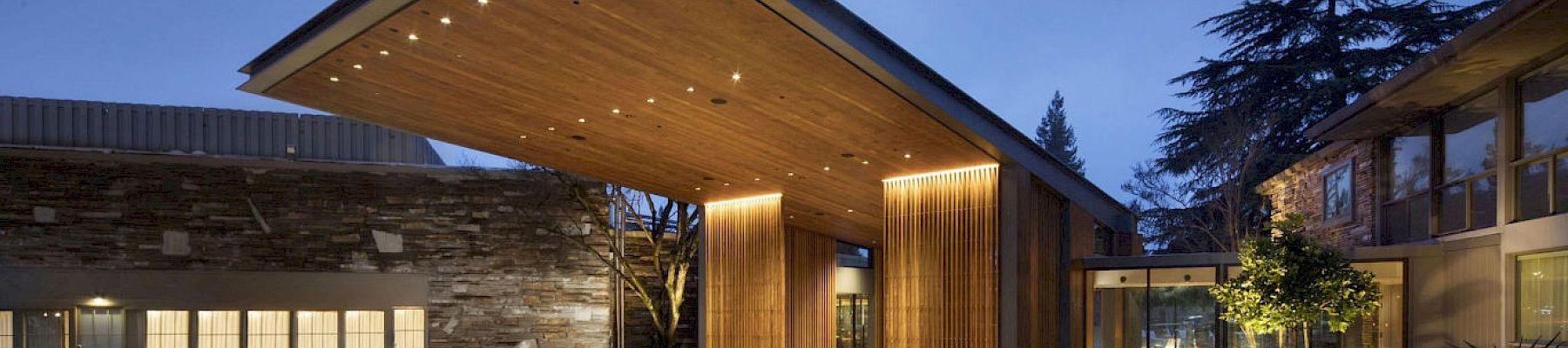 This image shows a modern building entrance with a large, illuminated wooden canopy and landscaped surroundings in the evening.