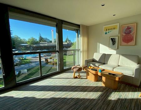 A bright, modern living room with a large window, couch, coffee table, wall art, and a view of an outdoor area with trees and a pool.