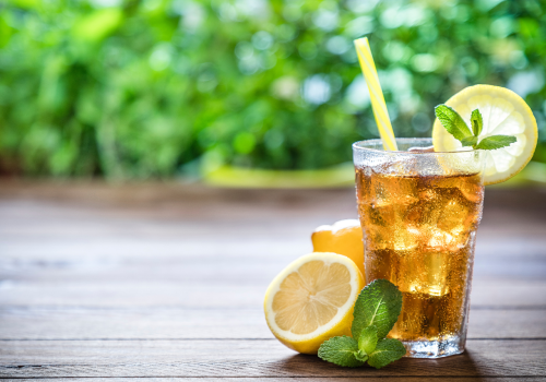 The image shows a refreshing iced tea with lemon slices and mint leaves on a wooden table, accompanied by a yellow straw, and a green, blurred background.