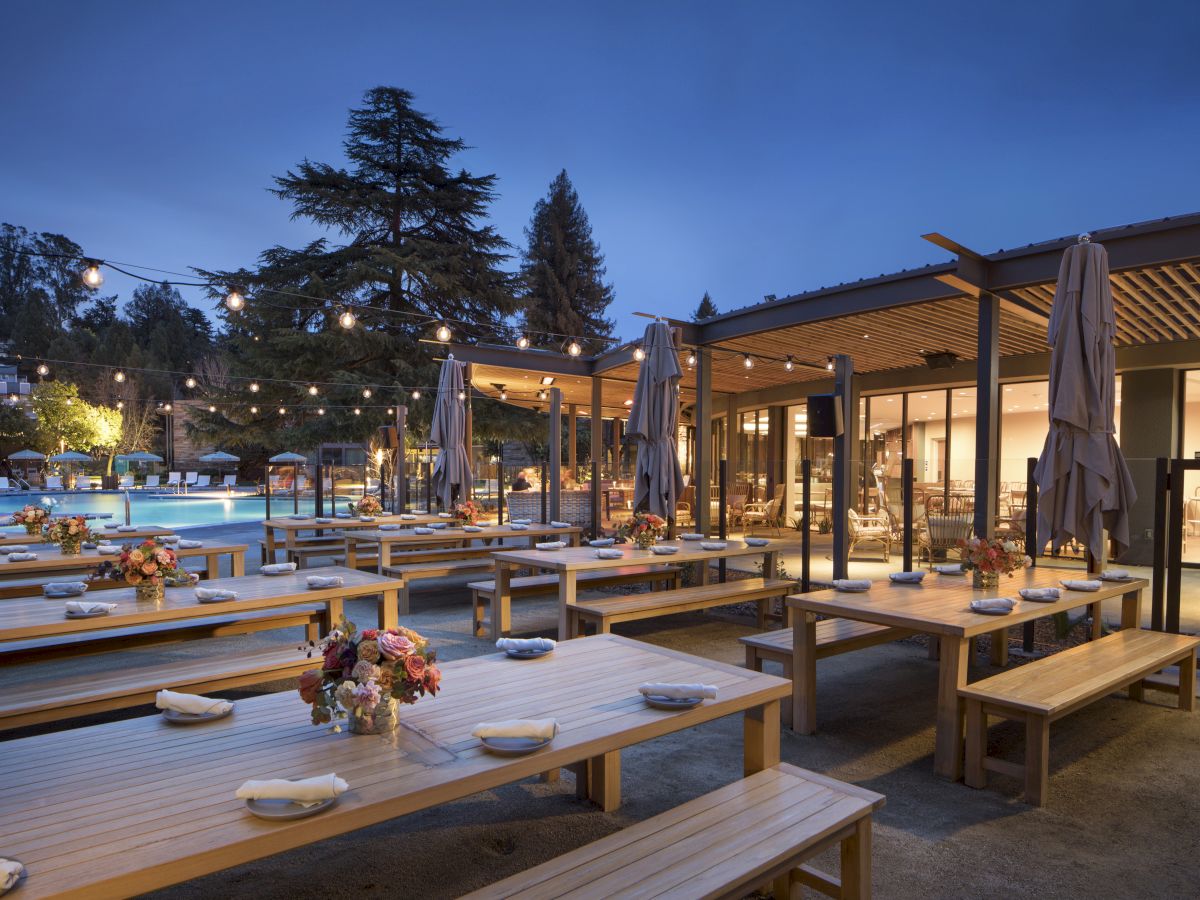 An outdoor dining area with wooden tables, benches, string lights, and floral centerpieces, near a building with large windows and a view of trees.