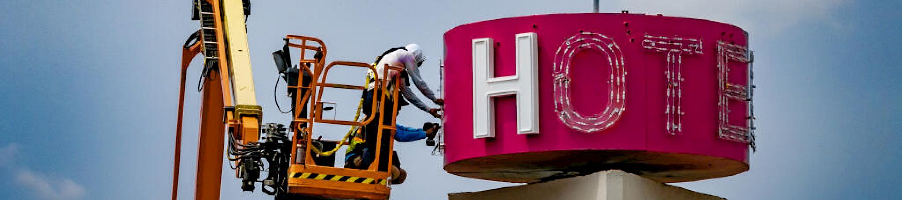 A worker on a lift is repairing a pink neon hotel sign featuring a flamingo against a backdrop of a partly cloudy sky.