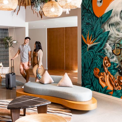 A modern lobby with a couple, luggage, stylish furniture, and a vibrant mural featuring a person with glasses, surrounded by nature and holding tea cups.