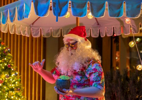 A person dressed as tropical-themed Santa with a red hat and floral shirt holds a drink under an outdoor umbrella, near a lit Christmas tree.